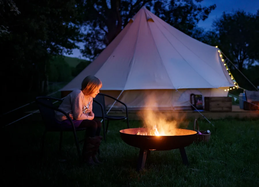 Girl watching the campfire with a bell tent in the background at Chalke Valley
