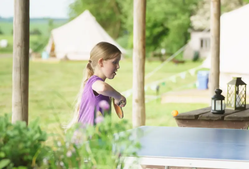 Girl plays ping pong in shelter with bell tent visible