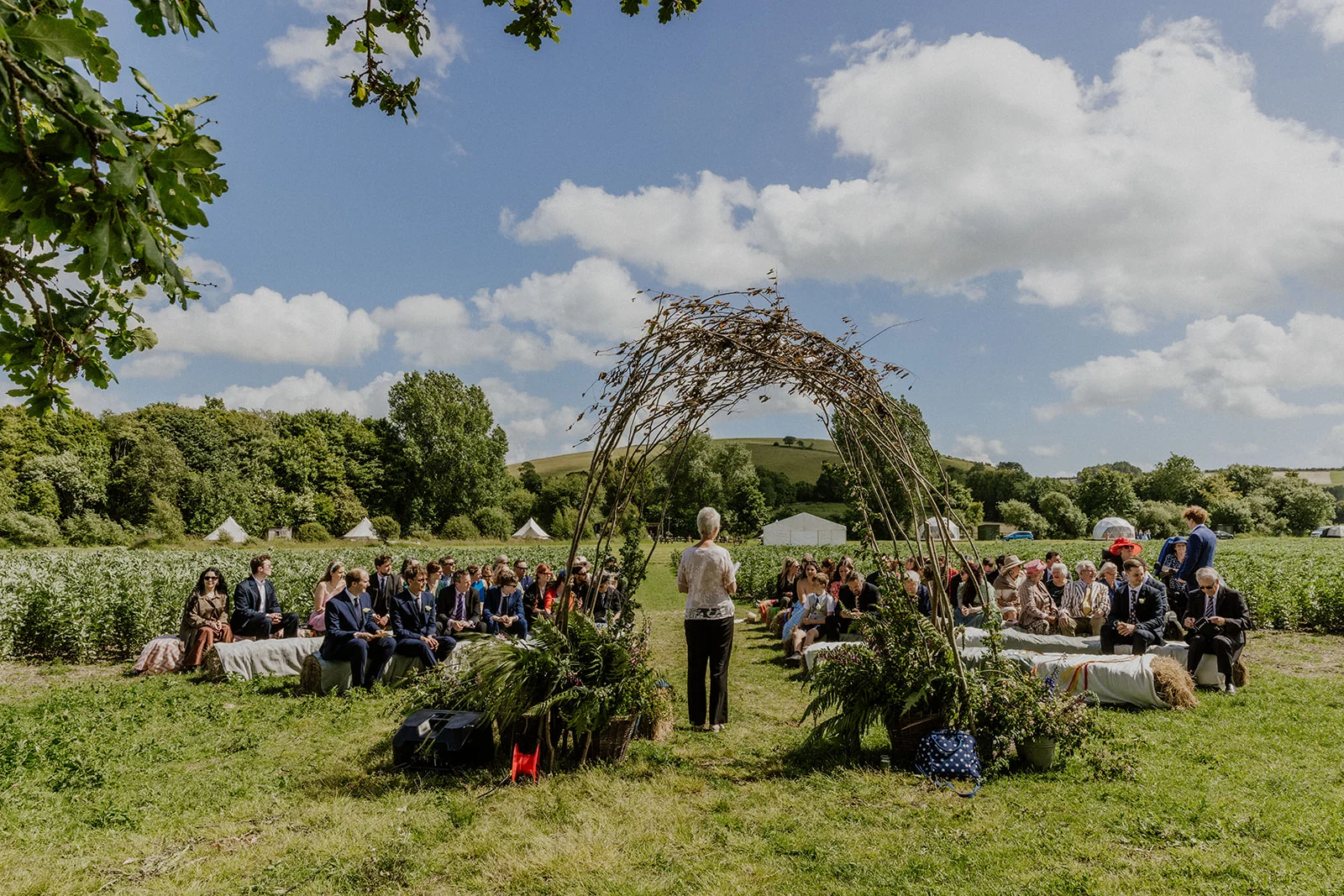 Wide shot of an outdoor glamping wedding with guests on straw bales at Chalke Valley